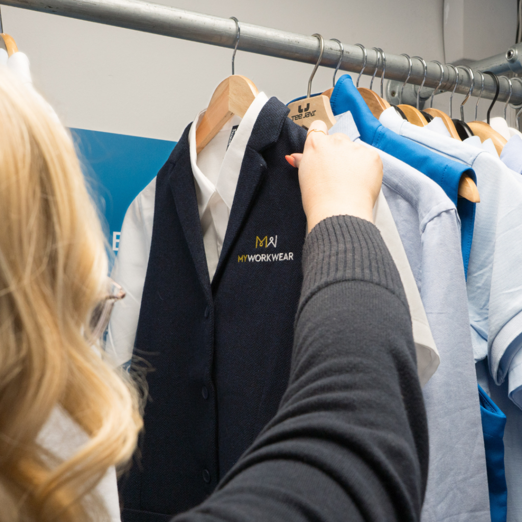 Back of womans head as she reaches to pick up a white shirt and blue overcoat off a rail, the clothing is branded with MyWorkwear.